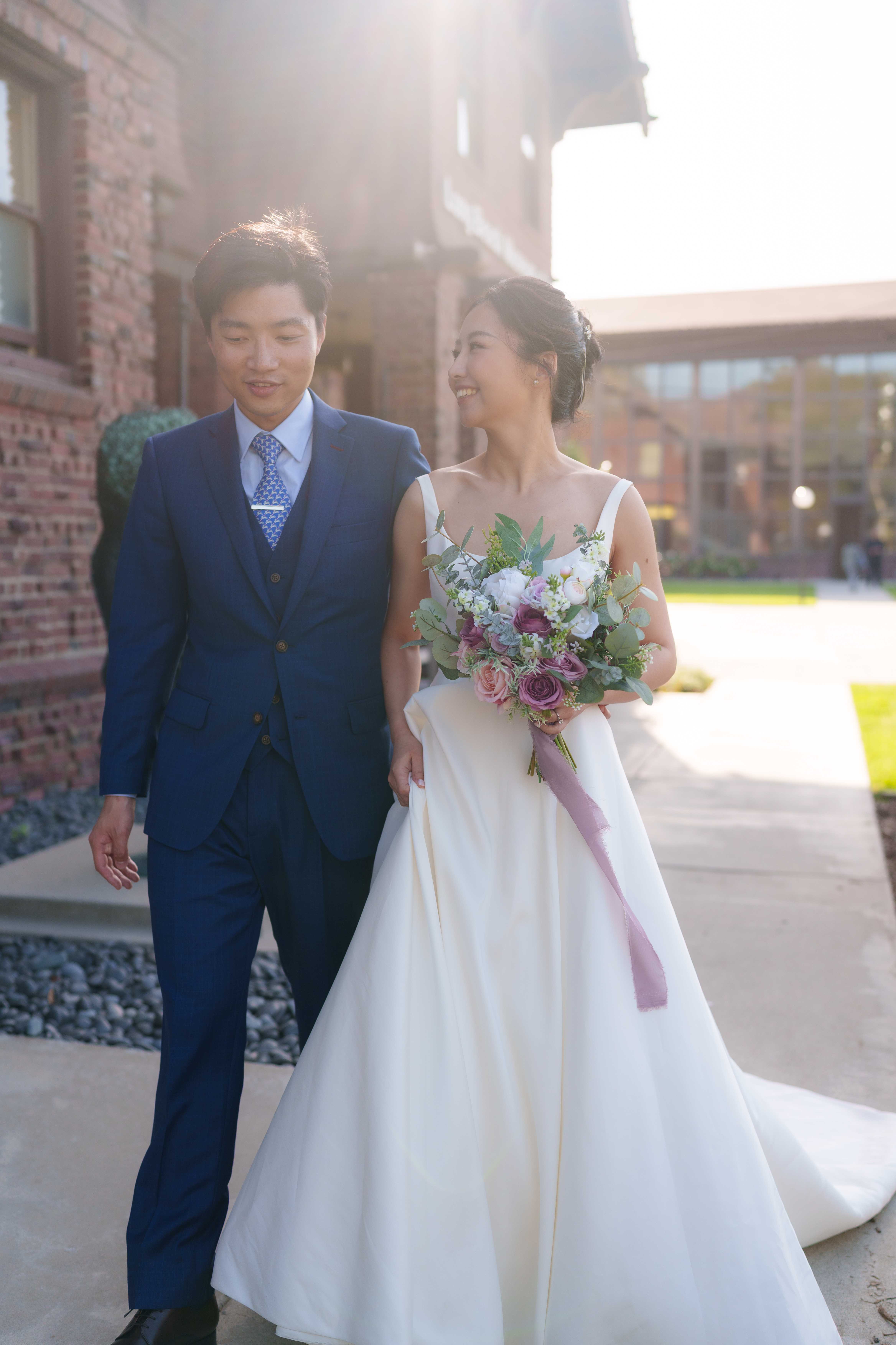 Newlyweds walking together outside a brick building in golden backlight, bride in a white gown with a floral bouquet