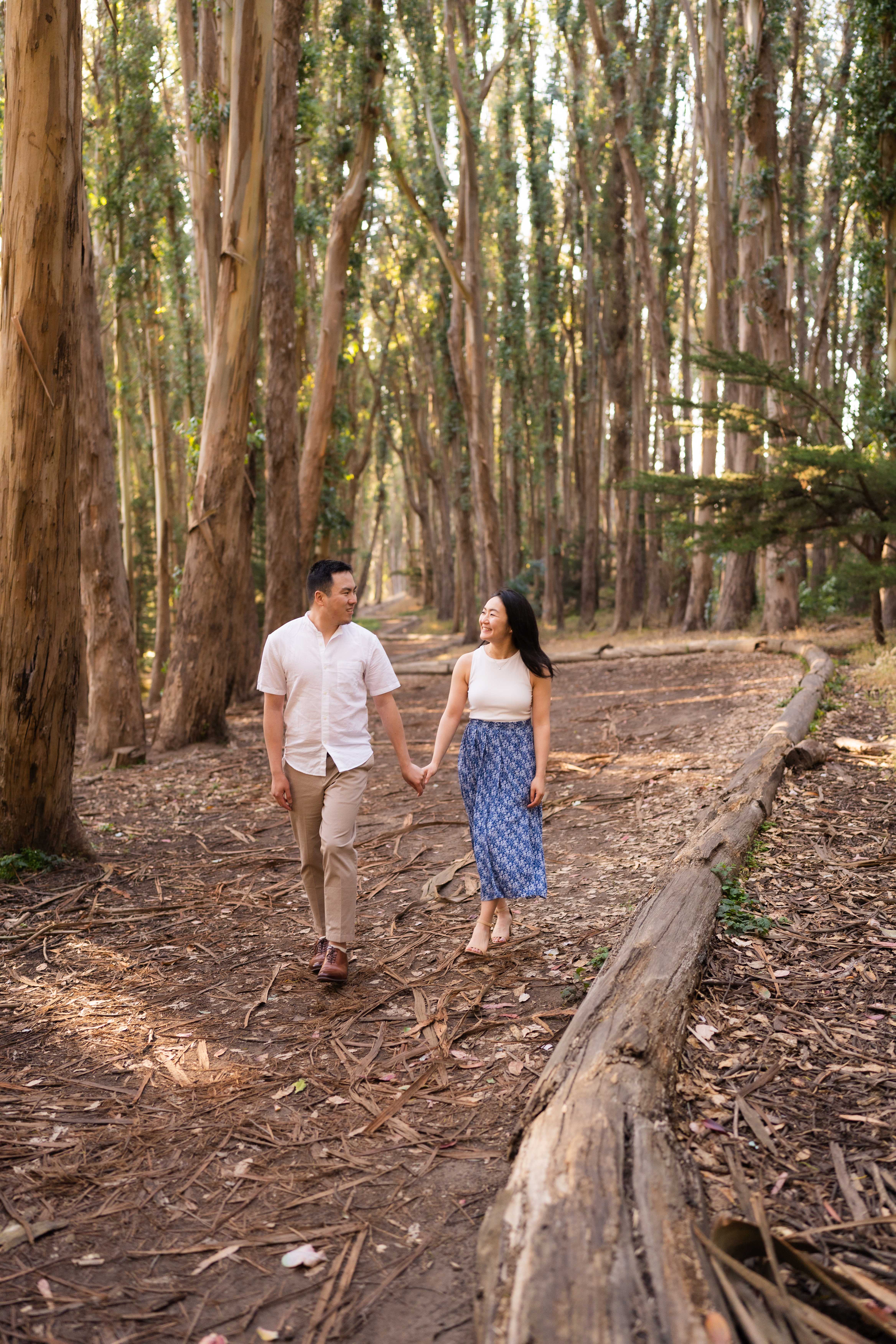 Couple walking hand in hand through a sunlit eucalyptus forest path, looking back at each other